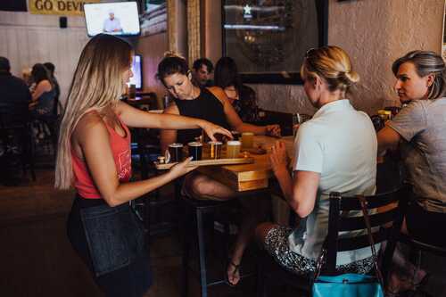 Three women sitting at table in restaurant being served beer flight sampler by server